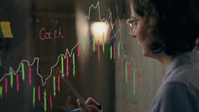 A top manager draws a business plan diagram on a transparent glass board in stylish conference room. Business data analyst looks for an error in the data after month of work using modern technologies