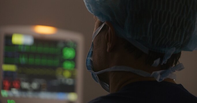 Close up of male surgeon in surgical suit using endoscope in surgery, looking at monitor. Electrocardiography screen shows condition of patient in the background. Modern equipment in operating room.