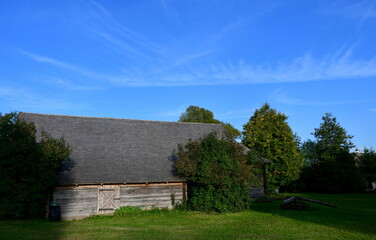 A view of an old wooden hut, shack, or stable surrounded with growing flowers, herbs, a wooden fence, and ancient farming equipment seen next to a well maintained lawn, pastureland, or meadow