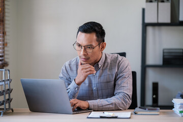 Portrait of young asian businessman, man smiling while sitting inside office, boss in business suit at workplace using laptop.