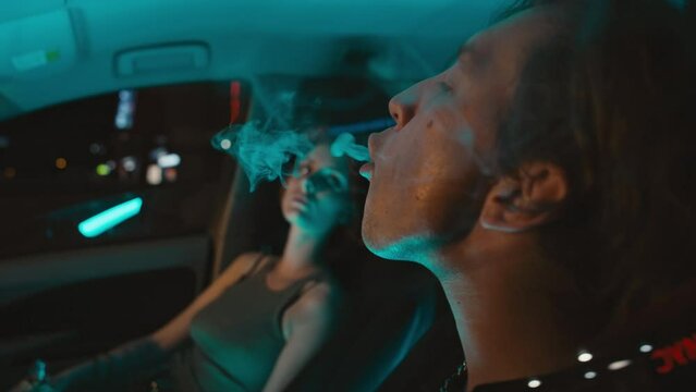 Close up side handheld shot of young Caucasian man sitting in parked car with girlfriend and exhaling vapour uncontented