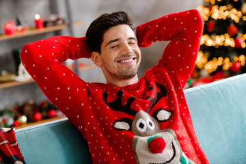 Photo of cheerful carefree young man sit cozy couch closed eyes hands behind head christmastime magic house inside