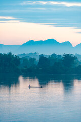 Silhouette of a fishing boat in the Mekong River, bordering Thailand and Laos, in the morning. The background is mountains. There is soft yellow sunlight shining, vertical image suitable for postcards