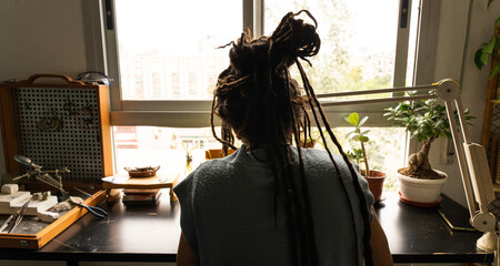 Boy with dreadlocks working in a goldsmith shop, designing jewelry pieces. Sitting at a table in front of a window.