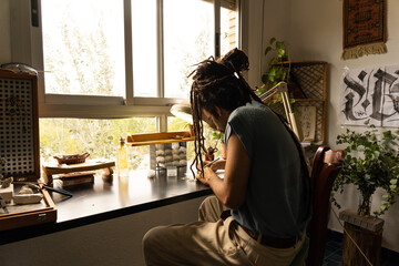 Young goldsmith working in his home workshop. Sitting in front of a window and a wooden table concentrating on his work. Rastafari
