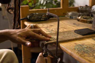 Hands of an artisan jeweler sawing a piece of jewelry with a hacksaw on a wooden board on a...