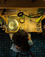 Birds Eye View of a goldsmith at a jeweler's bench working on handcrafted pieces. Young boy with dreadlocks