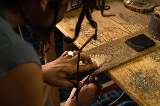 Young Rastafarian artisan jeweler cutting out a piece with a hacksaw on a table in the jewelry workshop. Copy space on the right