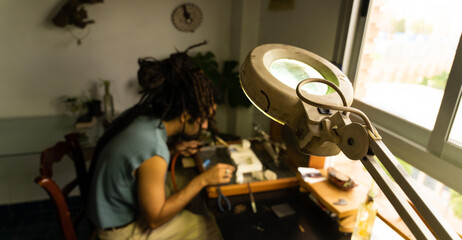 Lamp with focused magnifying glass. Behind a young goldsmith working on a piece of jewelry. Copy space to the right