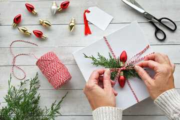 Woman wrapping Christmas gifts with red & white bakers twine, greenery & red light bulbs, flat lay