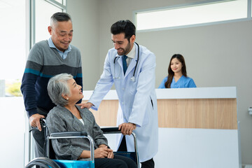 Obraz premium Selective focus of a smiling Caucasian male doctor in a white gown, standing holding a shoulder while talking to a senior Asian female patient sitting on a wheelchair pushed by her husband at hospital