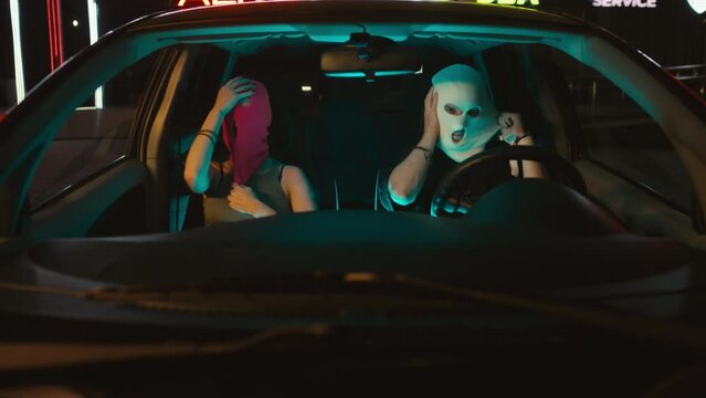 Medium handheld shot of young Caucasian couple of bandits high-fiving, putting balaclavas, taking guns, kissing and getting out of car at parking area at night