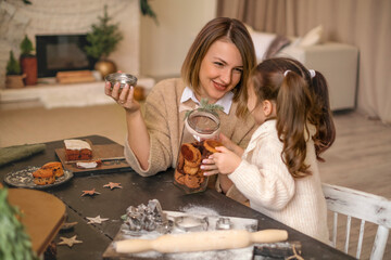 A mother treats her daughter to homemade gingerbread Christmas cakes from a jar sitting at a table...