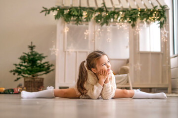 A thoughtful gymnast girl is sitting in a twine pose in a room decorated for the new year and looks away - obrazy, fototapety, plakaty