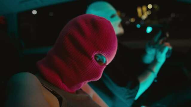 Close up side handheld shot of young female Caucasian bandit putting on balaclava, taking handgun and kissing her boyfriend in parked car at night before robbery