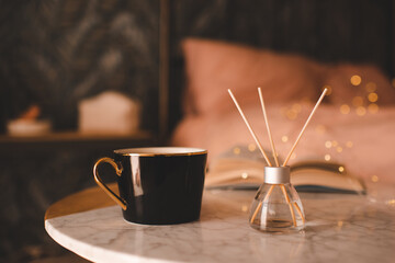 Cup of coffee with home liquid diffuser and open book on marble table in bedroom over glow lights. Hygge cozy home atmosphere. Aromatherapy. - Powered by Adobe