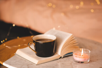 Mug of coffee on open paper book with burning scented candle on marble table over Christmas lights...
