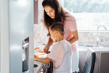 Loving young mother teaching her preschooler son prepare healthy food or salad at home kitchen, caring happy mom cooking together with kid on modern kitchen, boy involved in preparing food with mum
