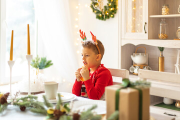 Cheerful little boy wearing warm red xmas sweater, deer horns on head eat dry orange slices in the...