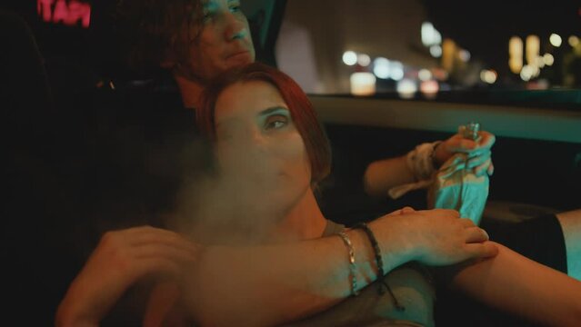 Medium arc handheld shot of young Caucasian loving couple hugging while drinking alcohol and vaping in parked car at night