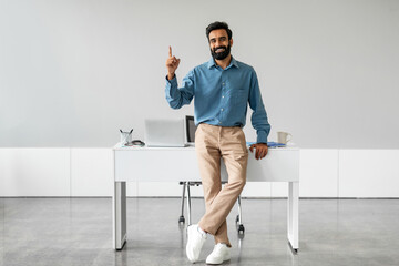 Handsome indian businessman sitting leaning on desk at workplace and pointing finger up at free place, smiling at camera - obrazy, fototapety, plakaty