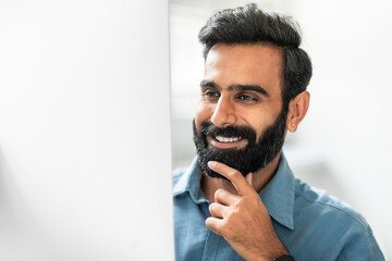 Happy indian businessman working on computer in office, looking at monitor and smiling, touching chin, closeup shot - obrazy, fototapety, plakaty