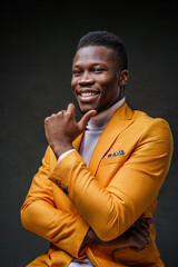 A joyful, dark-skinned man in a yellow jacket laughs while propping his chin with his finger against a dark background