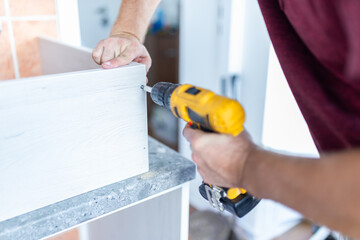 Carpenter installing kitchen chest of drawers by using electrical screwdriver. Home renovation concept.