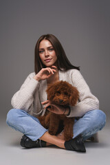 Attractive young brunette in white top and denim shorts, seated on the floor, cradling her brown toy poodle, against a gray studio backdrop