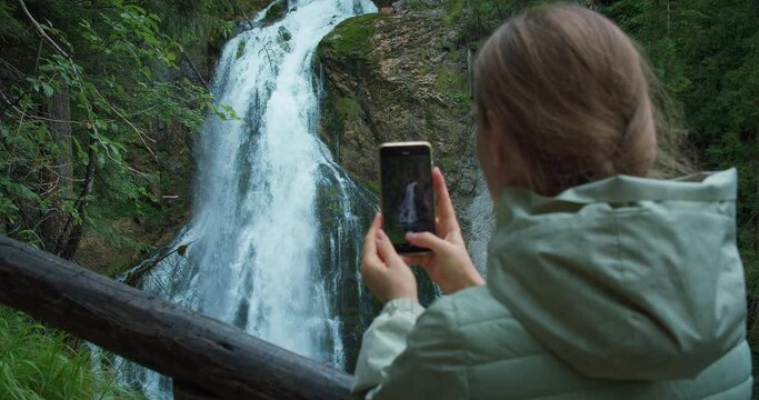 Woman takes a photo of a waterfall on phone. Girl travels in Austria. Tourist films Golling Wasserfall in mountain forest.