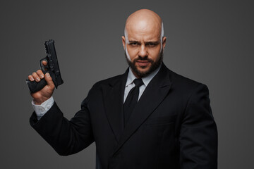 Bald security man in a suit holds a gun, ready to protect, against a gray studio backdrop
