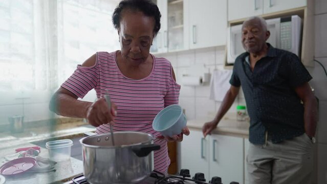 Elderly senior couple hanging in kitchen, an older black woman serving food into bowl while husband leans in background interacting with wife