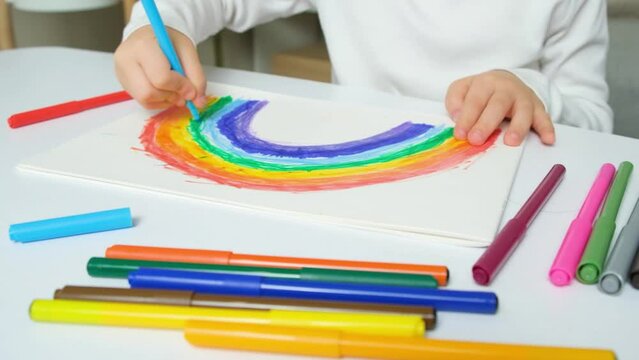 A small child draws a rainbow with felt-tip pens in a sketchbook while sitting at a table
