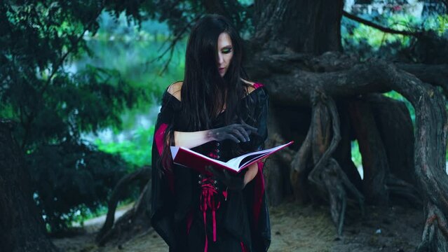 Fantasy woman sexy witch with long flowing hair holds old book reads magic spell. Girl fairytale goth vampire. Black red dress halloween gothic costume. Dark deep forest tree. Hands in black paint 4k