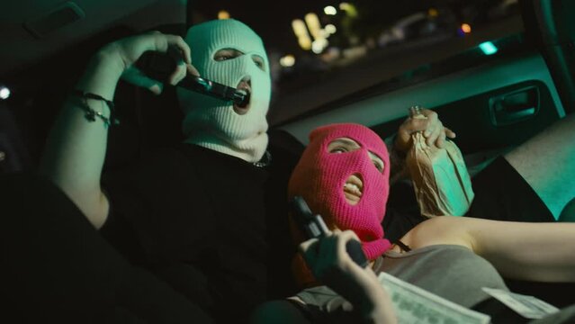 Medium handheld shot of young Caucasian robbers posing with handguns and taking selfie with flash lying in parked car with stolen money at night