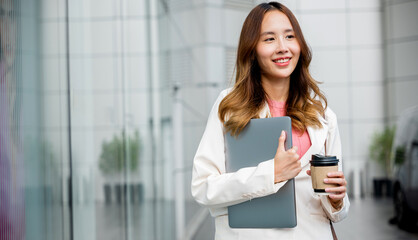 Business woman wearing white suit jacket and smile while go to work outdoor mirror building background. Confident Businesswoman with a cup of coffee and holding Laptop walking outside office building