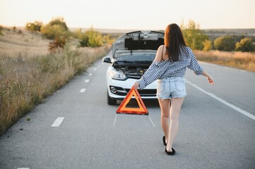 Beautiful sexy woman holding an emergency stop sign near a broken car. - obrazy, fototapety, plakaty