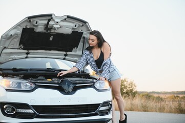 Attractive slim young girl in summer shorts and shirt repairs a broken car. A beautiful woman stands near raised car hood.