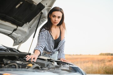 Beautiful slim girl in shirt and shorts looks in open car hood on a road