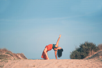 Young girl on the beach sand doing a flexible yoga pose with a blue sky in the background.