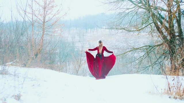 Fantasy happy woman standing dancing on top of mountain holds skirt dress in hands, red fabric flies waves in wind. Princess girl enjoys winter nature fresh cold white snow. Lady back rear view art 4k