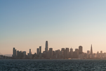 Naklejka premium A picturesque skyline of San Francisco Panorama at sunset golden hour from Treasure Island, California, United States. Panoramic view of cityscape with mist and foggy air. Illuminated dusk city