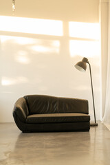 A black sofa and a floor lamp on the background of a white wall. Light from the sunset on the wall.