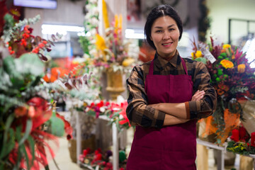 Positive asian female flower shop seller in uniform standing with crossed arms near flowers