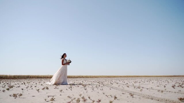 Pretty young bride holding delicate bouquet at desert landscape, windy weather. Elegance lady waiting for a groom after wedding ceremony. Attractive model posing in gorgeous white dress in nature.