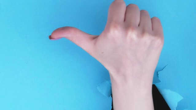 Vertical video. Dislike gesture. Negative feedback. Female hand showing refusal thumb down in breakthrough paper hole on blue torn wall background with copy space set of 2 loop.