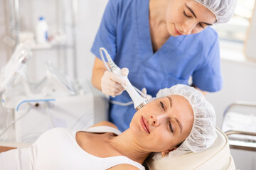 Naklejka premium Young female cosmetologist performs hardware facial ultrasound procedure to young female patient