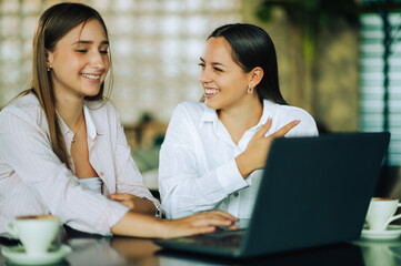 Two happy female friends are sitting in a coffee shop and searching the internet