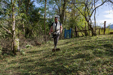 A man uses a trimmer to mow the grass in the backyard of his rural home, located in an orchard, on a sunny autumn day. Highlighting the concept of maintaining a countryside estate
