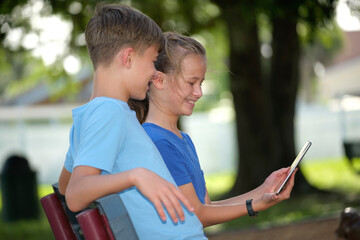Two happy teenager children, girl and boy looking at screen of digital tablet, reading, studying or gaming together sitting on bench outdoors on summer sunny day